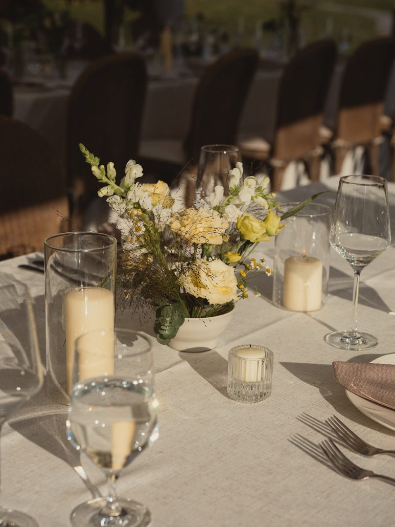 Close up of soft yellow and white floral centerpieces with candles and glassware catching the afternoon light