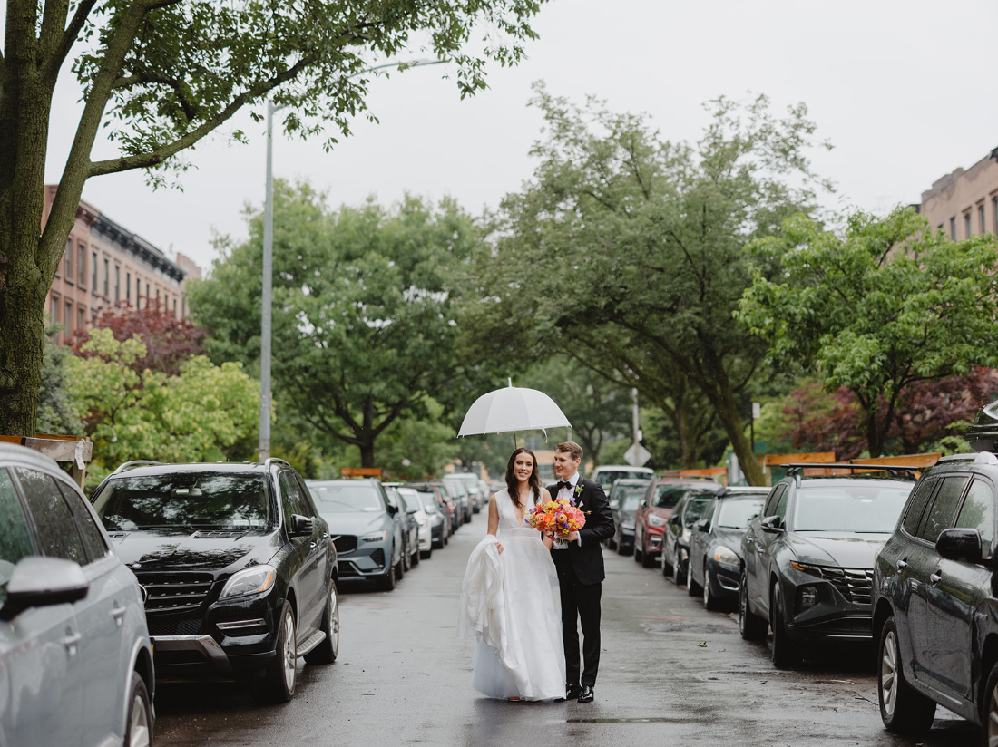 Newlywed couple walking under an umbrella on a rainy city street during their wedding weekend celebration.