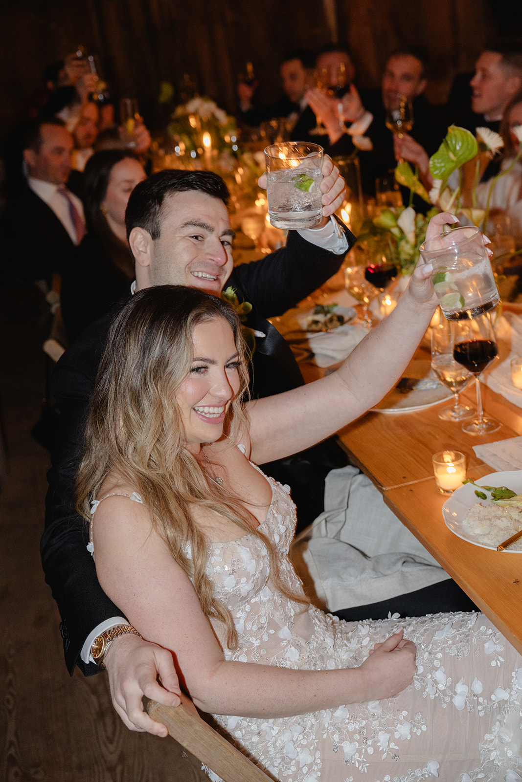 Couple toasting with guests at a candlelit reception dinner inside a rustic wooden barn.