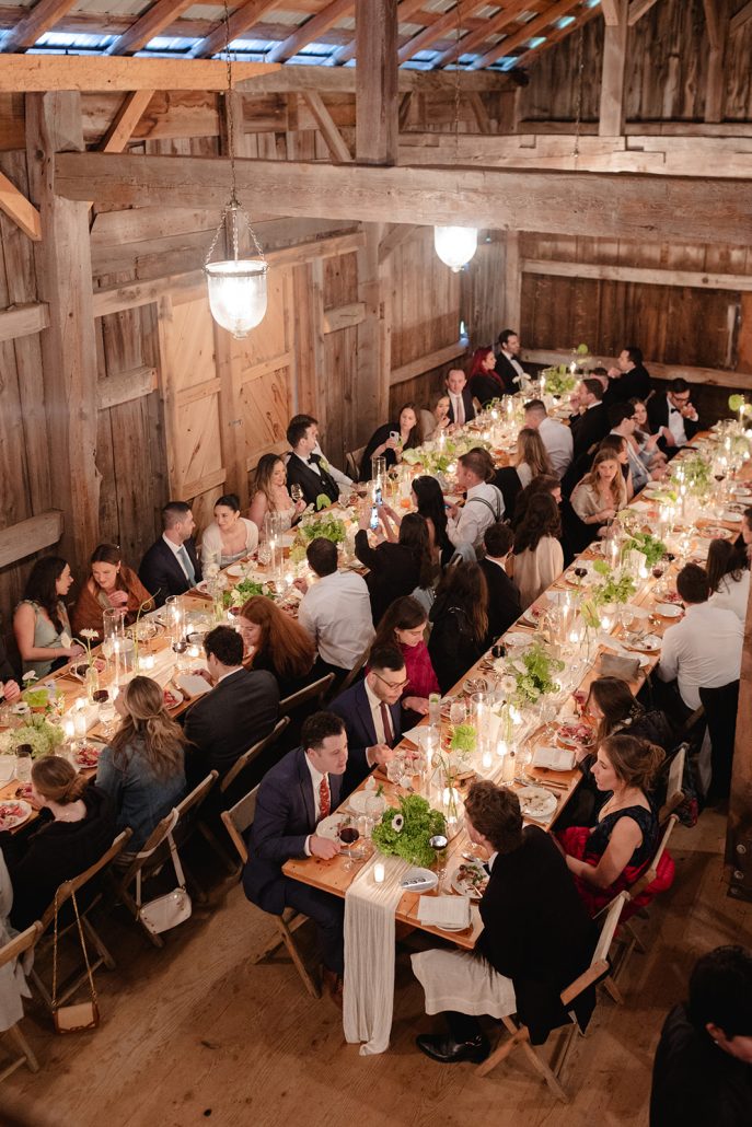 Guests seated at long farmhouse tables under warm candlelight at a wedding weekend welcome dinner.