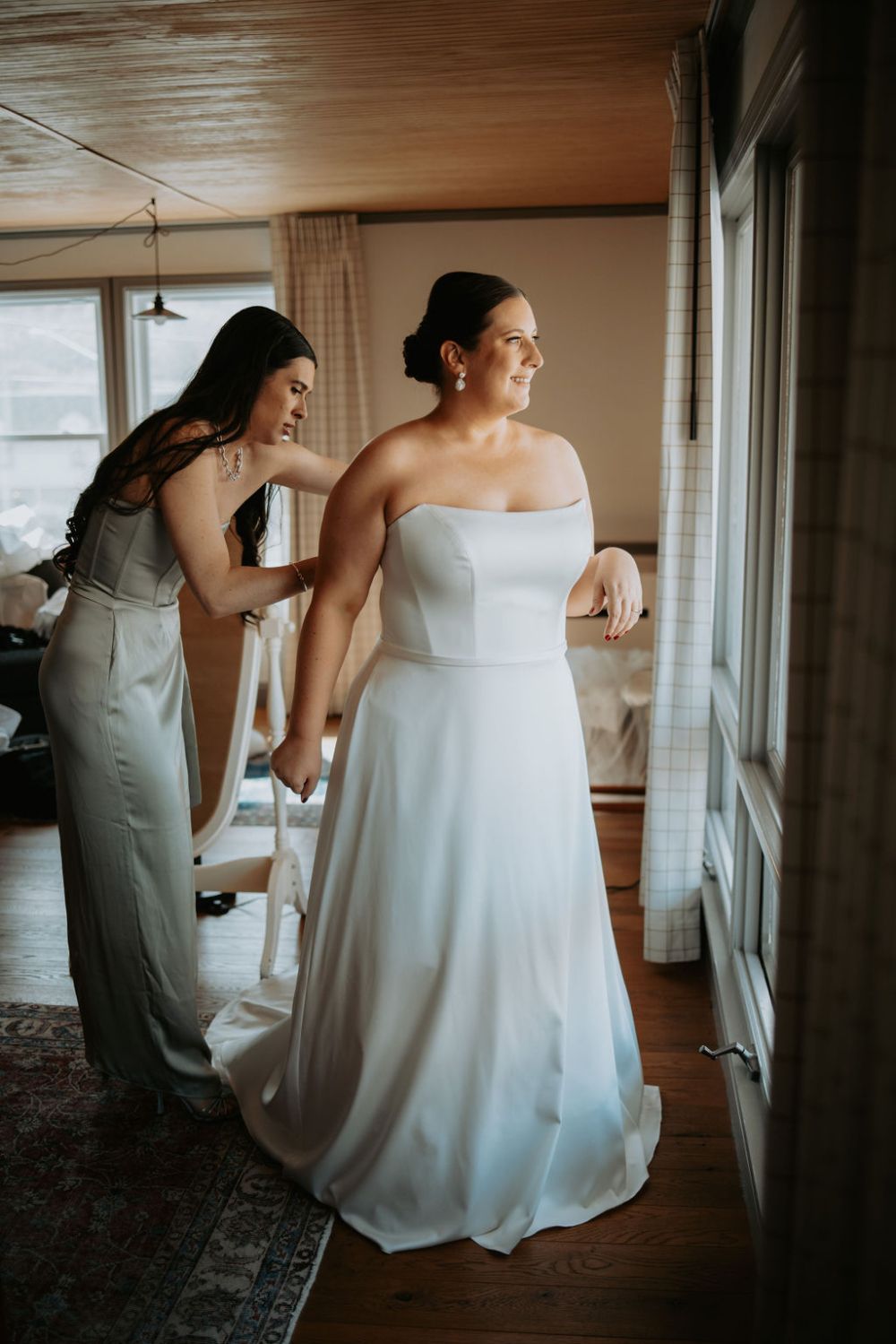 Bride standing by the window while a bridesmaid fastens her gown during wedding morning preparations