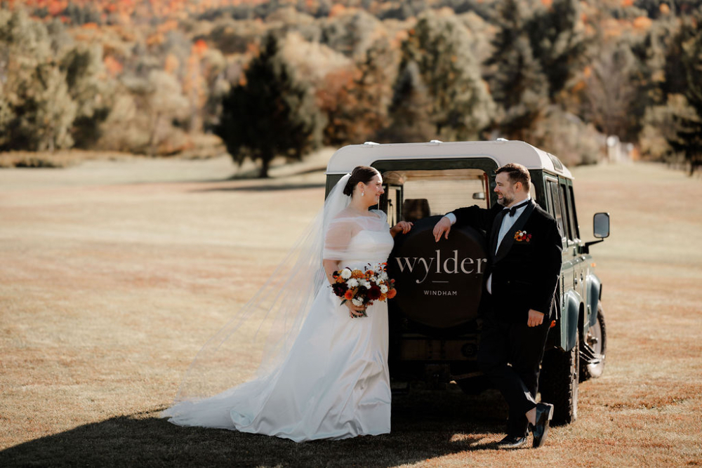 Bride and groom posing beside a vintage vehicle at their Hudson Valley venue, reflecting how much does a luxury wedding cost through fashion, setting, and photography