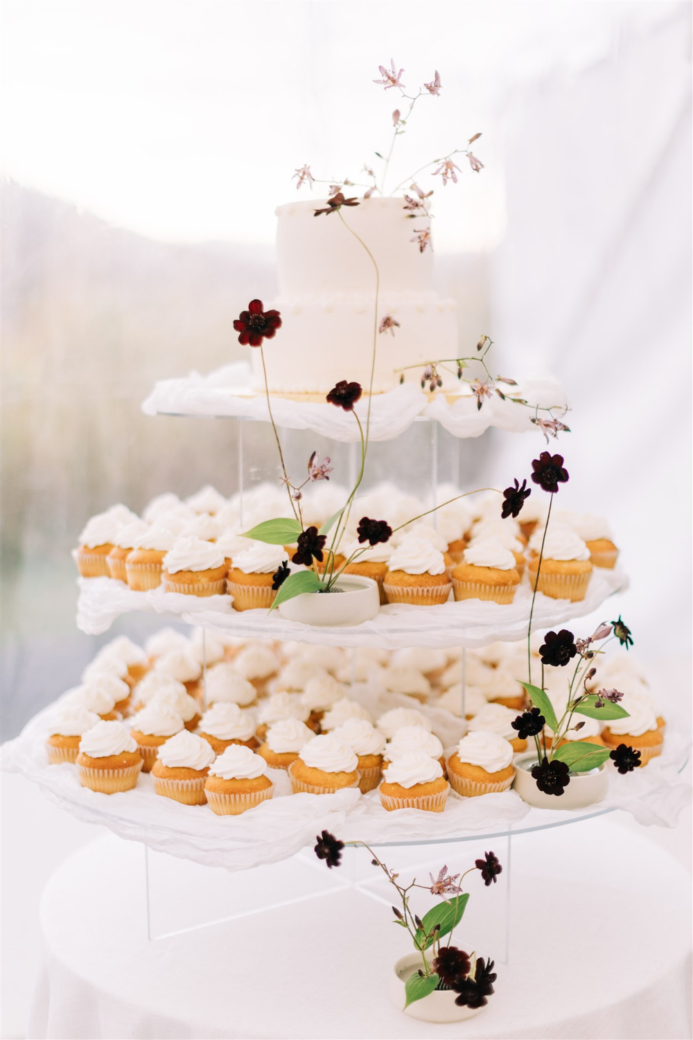 Tiered wedding cake surrounded by cupcakes and minimalist floral accents