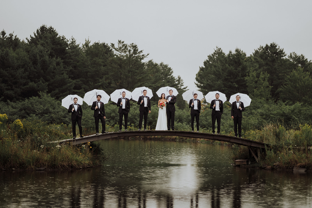 Wedding party standing on a wooden bridge holding umbrellas over a serene pond, coordinated with wedding planner responsibilities