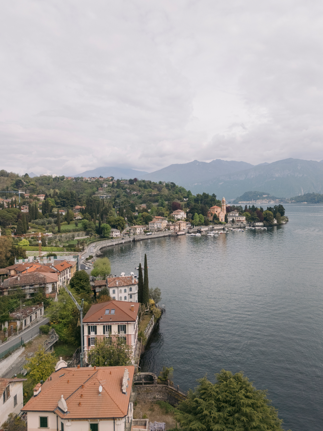 Scenic aerial view of the Lake Como waterfront, historic church, and hillside villas beneath dramatic clouds.
