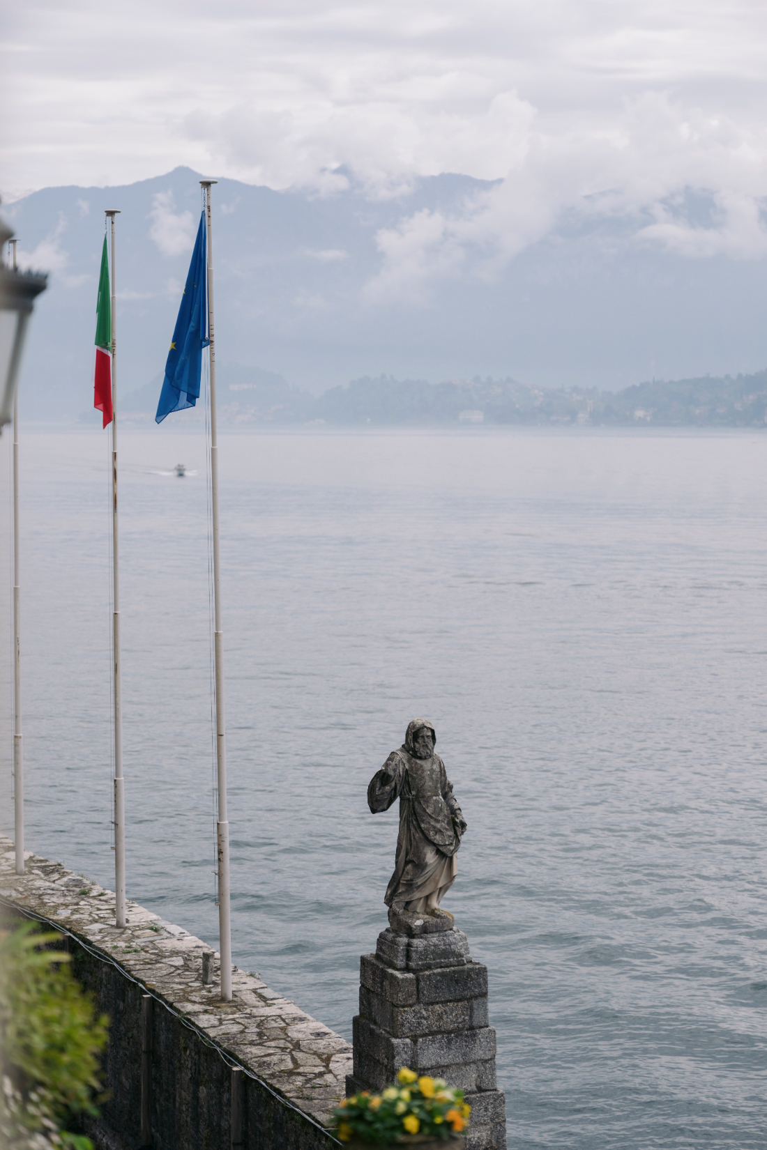 Historic stone statue and Italian flags overlooking Lake Como from the waterfront terrace at a Villa del Balbianello wedding.
