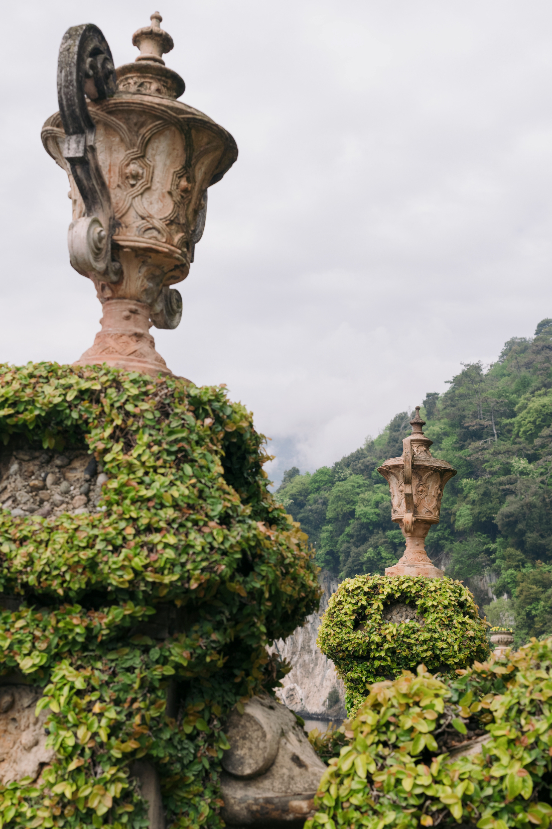 Ornate stone urns surrounded by manicured topiary with Lake Como and lush mountains in the background.