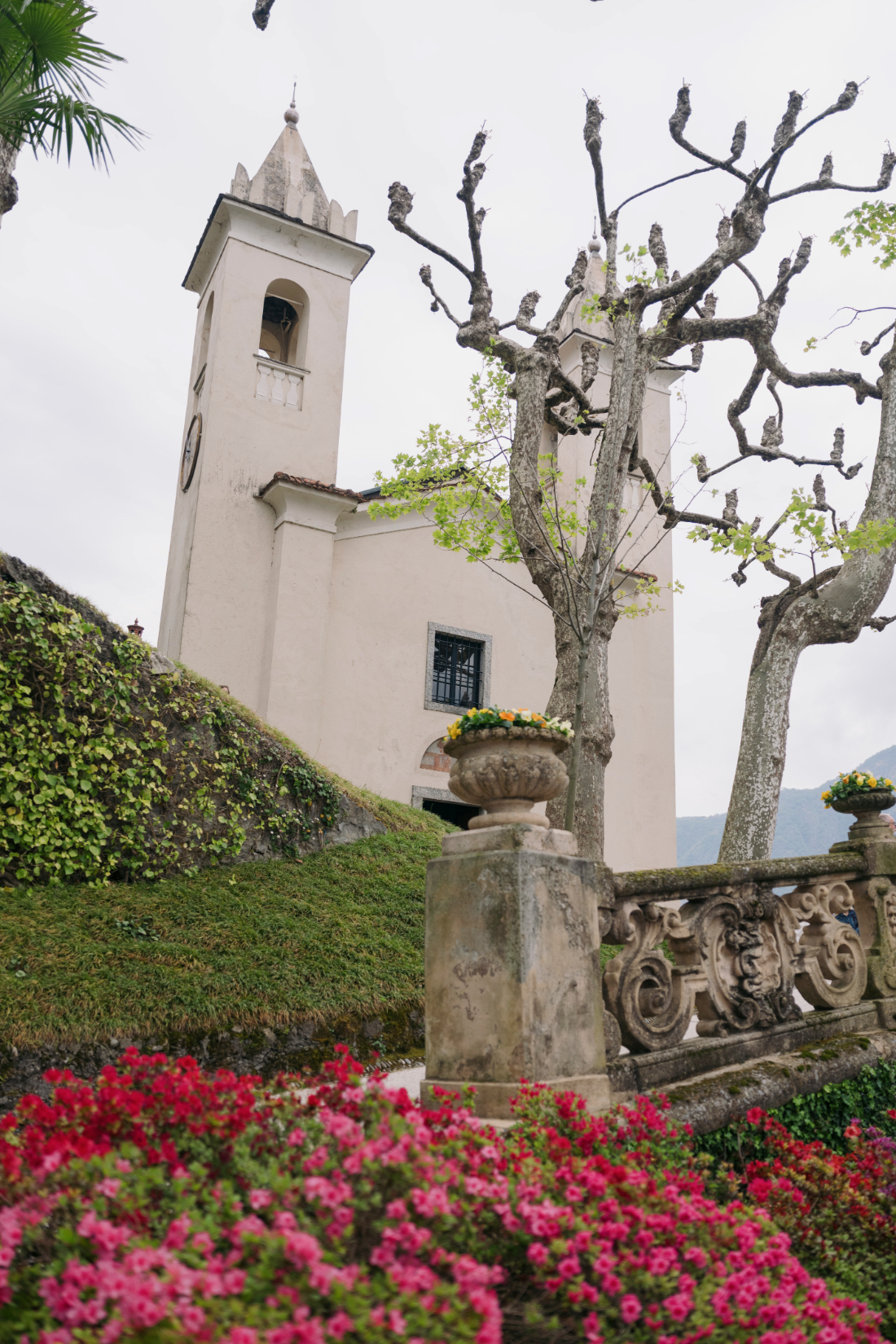 Historic church framed by vibrant spring flowers, stone balustrades, and sculpted trees overlooking Lake Como.