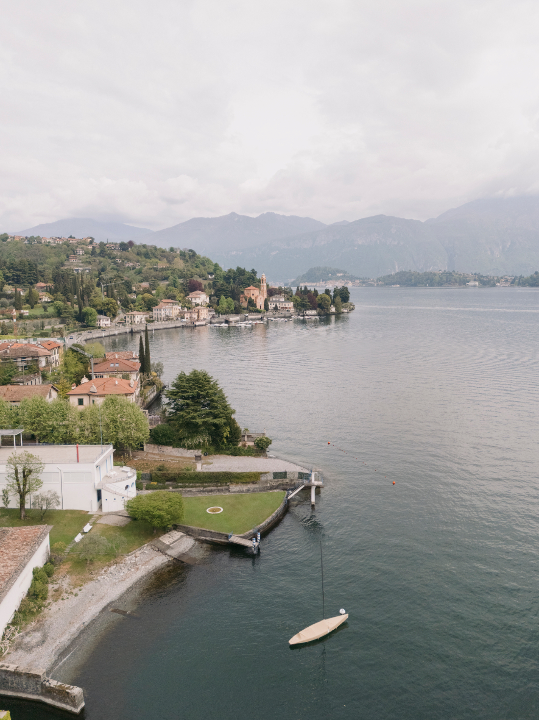 Panoramic aerial view of a charming Lake Como village, historic church, and tranquil waterfront surrounded by mountains.