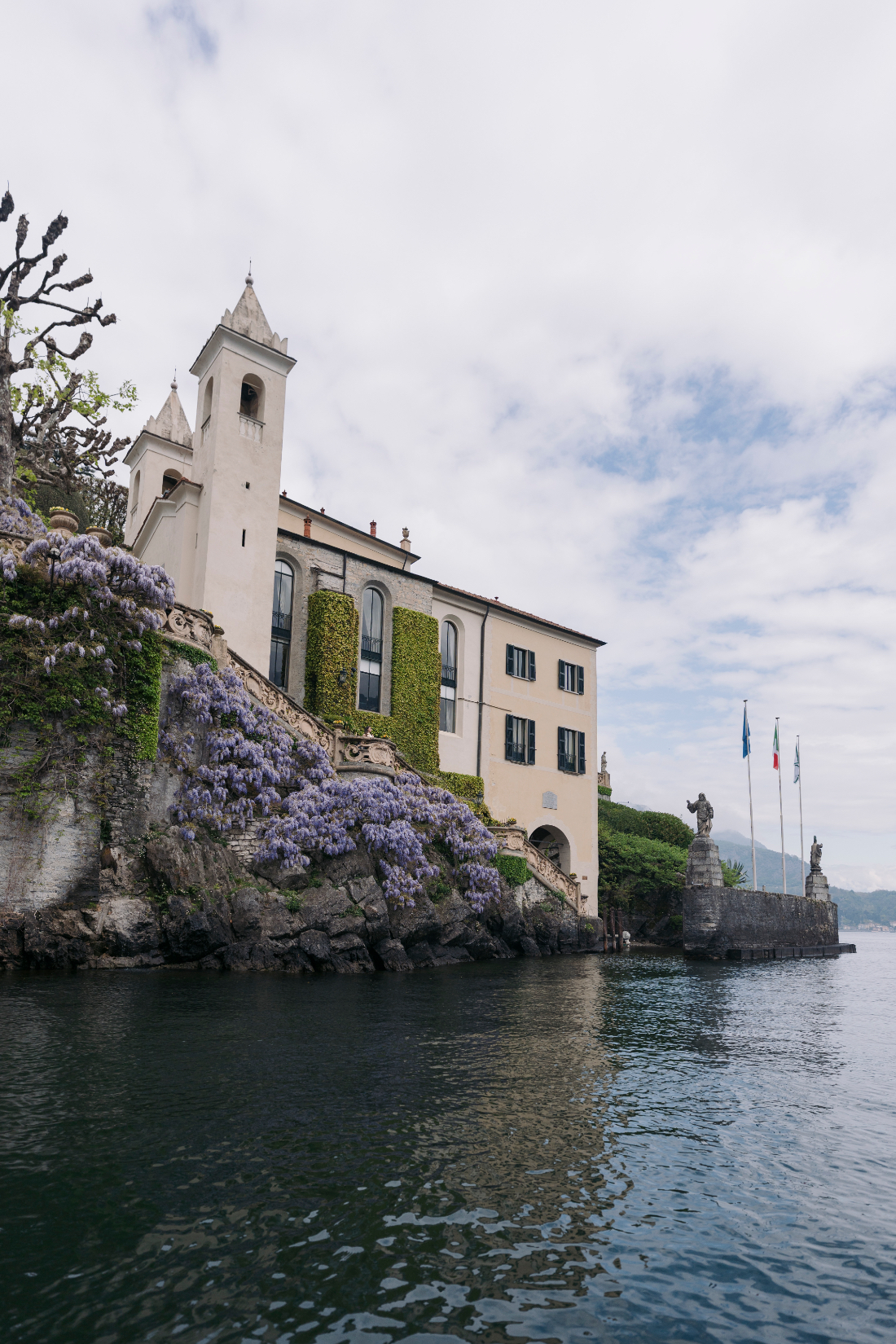 Historic bell tower, ivy-covered walls, and cascading purple wisteria overlooking the lake.