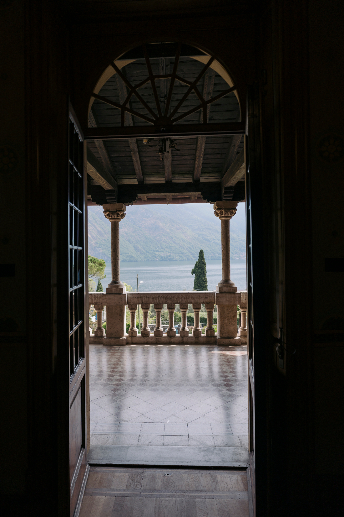 Framed view through open doors to a stone balcony overlooking the tranquil waters of Lake Como and surrounding mountains.