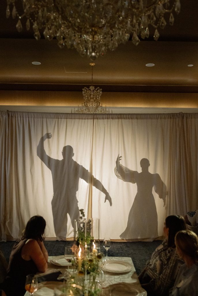Guests watch a dramatic shadow dance performance behind illuminated draping during an intimate candlelit dinner.